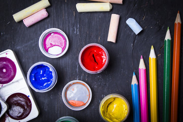 Bright multi-colored paint, pastels, crayons, pencils, brushes on a black wooden background. Set...