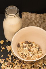 Bowl of muesli with dry fruits and bottle of plant milk, on black background.