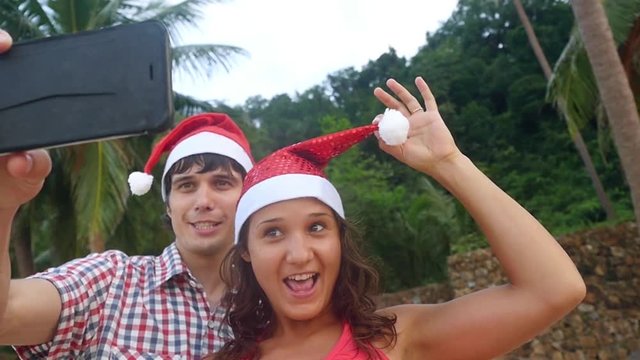 Happy beautiful couple on Christmas travel holidays taking selfie picture with smartphone wearing santa hat during sunset in slow motion. 1920x1080
