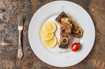 Fried fish fillet with lemon and tomatoes on a white plate. Old background. Top view. Close-up