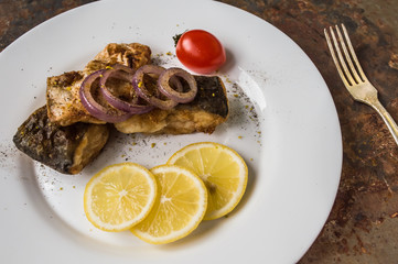 Fried fish fillet with lemon and tomatoes on a white plate. Old background. Top view. Close-up