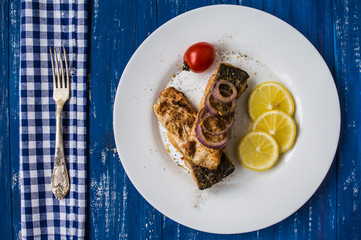Fried fish fillet with lemon and tomatoes on a white plate. Wooden blue background. Top view. Close-up