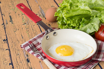 
Fried egg and vegetables.    Fried egg in a frying pan on a checkered napkin, near fresh green lettuce, red ripe tomatoes, raw egg and fork on old wooden table.