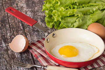 

Fried egg.    Fried egg in a frying pan on a checkered napkin, near fresh green lettuce, raw egg and fork on old wooden table.