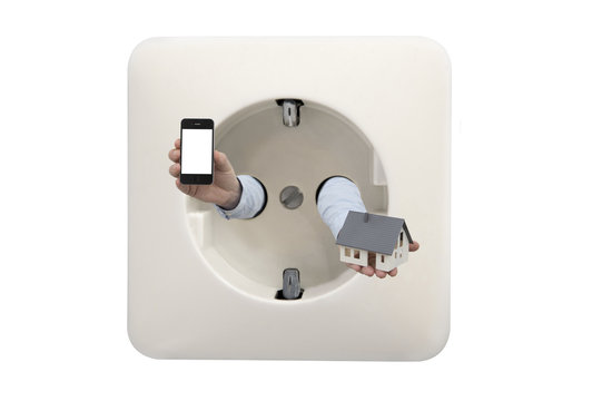 Close up on wall electrical outlet with mans hands holding smartphone and house model as concept for smart home app