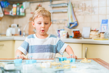 Fototapeta premium Little blond kid plays with molds for making ginger biscuits or cookies, sitting at the kitchen table with raw dough and wheat flour. Rapt face expression.