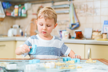 Fototapeta premium Little blond kid plays with molds for making ginger biscuits or cookies, sitting at the kitchen table with raw dough and wheat flour. Rapt face expression.