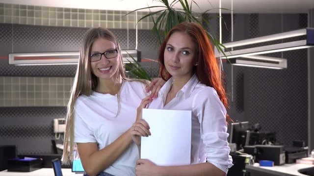 Two sexy office collaborators poses for the camera with a sheet of white paper