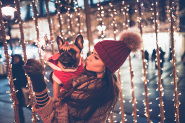 Beautiful young brunette woman with French bulldog enjoying Christmas or New Year night on a city...