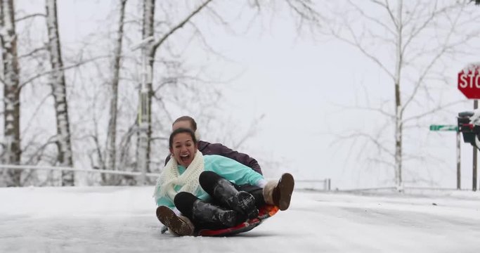 Two happy girls riding a snow sled down an icy hill in winter