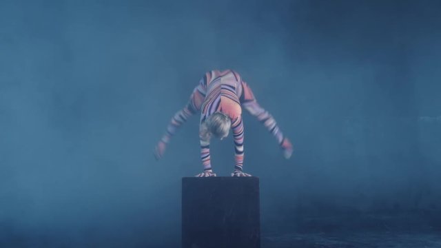 Young flexible blonde circus acrobat posing in studio in costume. Doing equilibre balance handstand on a cube. Copy space text.