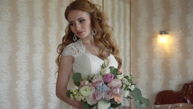 Young bride in white wedding dress holds a bouquet with curly hair and lace veil, stands in a room with beige walls.