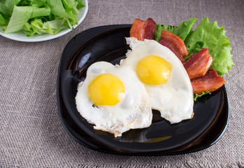 Black plate with fried eggs, bacon and lettuce closeup