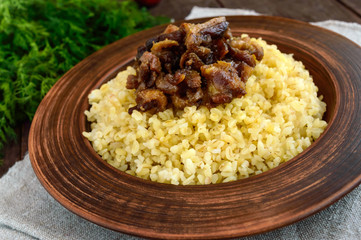 Eastern traditional wheat porridge - bulgur with roasted pieces fatty meats, bacon in a clay bowl on dark wooden background. Close up