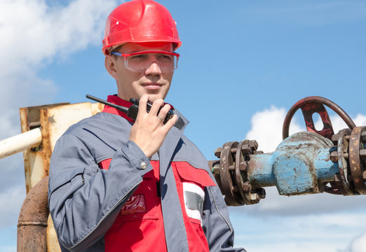 Worker Near Wellhead Valve Holding Radio And Wearing Red Helmet In The Oilfield. Oil And Gas Concept.