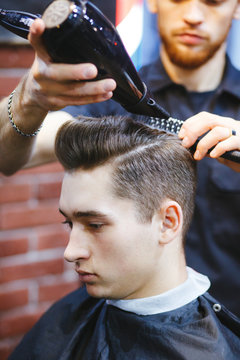 Male barber makes hair styling using a dryer.