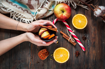 Female hands holding a hot mulled wine with spices, apple and orange on wood