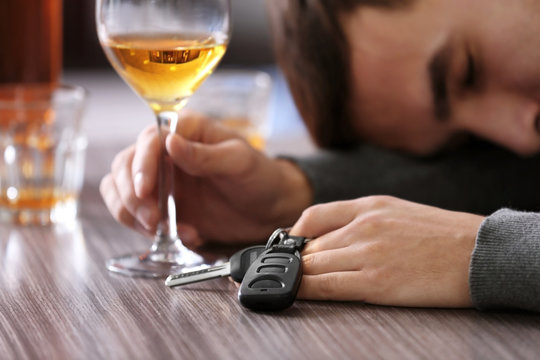 Drunk man sitting at table with car key and glass of alcoholic beverage, closeup. Don't drink and drive concept