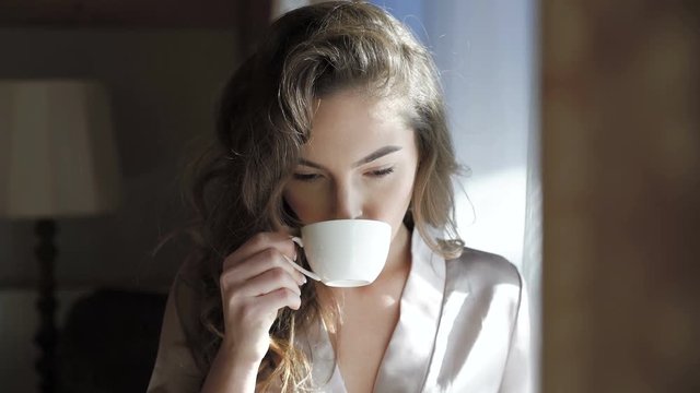 Pretty girl in nighty drinks a cup of coffee near the window in 4K