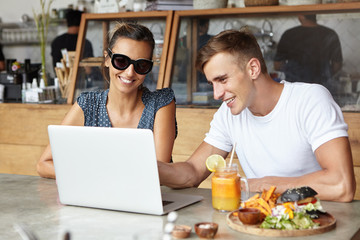 Handsome young man wearing white t-shirt showing something on laptop pc to his attractive female companion in stylish sunglasses during lunch at cafe, woman looking at screen with joyful smile