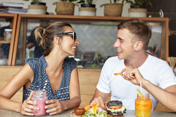 Happy couple having lively conversation on their first date, having joyful and carefree expressions, looking at each other and laughing. Man in white t-shirt sharing positive news with woman in shades