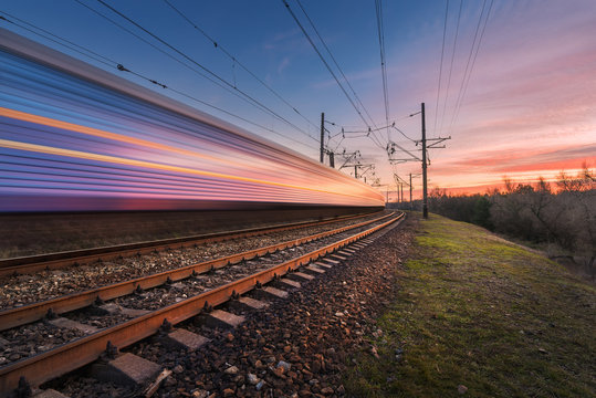 Fototapeta High speed passenger train in motion on railroad track at sunset. Blurred commuter train. Railway station against colorful sky. Railroad travel, railway tourism. Industrial landscape in the evening
