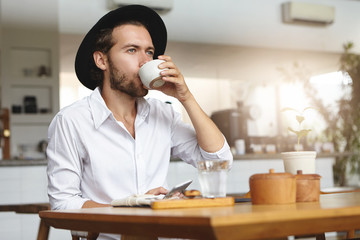 Fototapeta na wymiar Fashionable young man with beard wearing hat and white shirt having hot drink, sitting at table and holding gadget in his hand. Caucasian male using mobile phone, drinking tea or coffee at cozy cafe