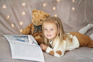 Fototapeta na wymiar little girl lying on the couch at home and reading a children's winter wonderland, teddy bear sits next