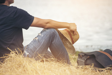 Smart man wear jeans to holding a hat, sitting on the golden grass,look forward to river.