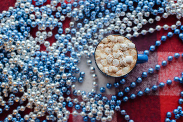 top view cup of cocoa with marshmallows near blue bead on red checkered background