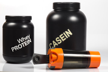 Whey protein black jar and big Casein protein black jar. Orange shaker lying near with opened cap. Close up image. White background. Shadows.