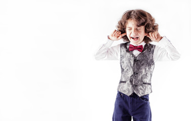 Curly cute boy in waistcoat and bow tie, his eyes shut, his fingers covering his ears and screaming. White background.
