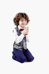 Cute cheerful curly-haired boy on his knees, hands clasped in prayer and looking into the camera. Close-up. Gray background.