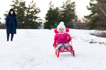 The child, a little girl riding on a sled with snow slides. Winter fun for children. A child with his mother and a winter walk.