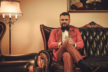 Extravagant stylish man with glass of whisky sitting on classic leather sofa in gentleman club