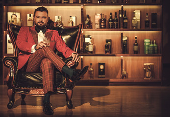 Extravagant stylish man with whisky glass sitting on armchair in gentleman club