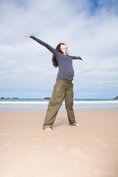 brunette brown hair pregnant woman with grey shirt green trousers and black sunglasses happy joyful open arms in sand beach ocean in Asturias Spain
