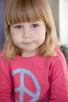 Portrait of three years old child face, blonde bang, with pink shirt blue peace hippie symbol, looking at camera seriously

