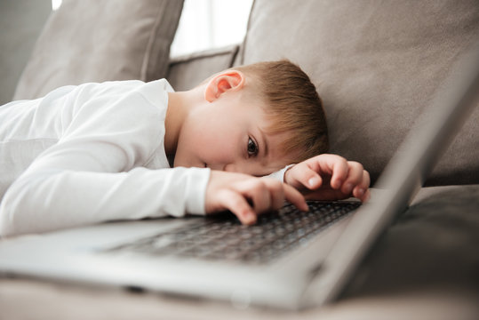 Little alone sad boy lies on sofa while using laptop