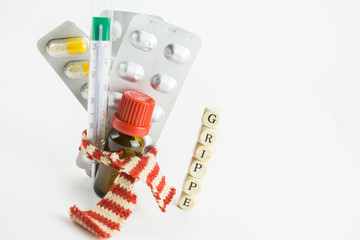 Dreierlei Tablettenblister, ein Fieberthermometer und ein Arzneifläschchen, gebündelt mit einem rot weiß gestreiften Schal, daneben das Wort Grippe mit Buchstabenwürfeln gelegt