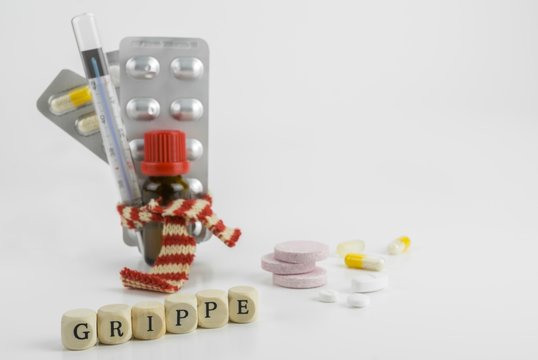 Viele Medikament, Tabletten und Hustentropfen, mit einem Schal gebündelt, daneben lose Tabletten und das Wort Grippe mit Buchstabenwürfeln gelegt