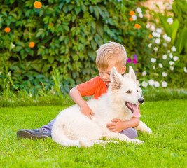 Young boy embracing White Swiss Shepherd`s puppy on green grass