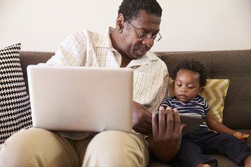 Grandfather And Grandson Sit On Sofa At Home Using Laptop