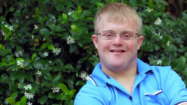 Close up face shot of friendly young man with down syndrome doing thumb up symbol against outdoor green background.