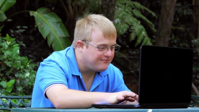 Close up of young man with down syndrome working on laptop in garden. Young man wearing eye wear doing thumbs up symbol.