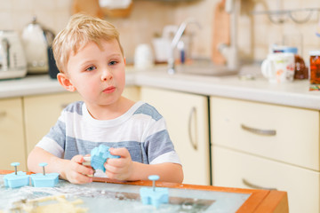 Little blond kid plays with molds for making ginger biscuits or cookies, sitting at the kitchen table with raw dough and wheat flour. Rapt face expression. Looking ahead.