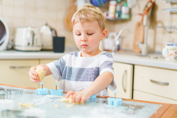 Little blond kid plays with molds for making ginger biscuits or cookies, sitting at the kitchen table with raw dough and wheat flour. Rapt face expression. Looking ahead.