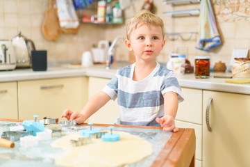 Fototapeta premium Little blond kid plays with molds for making ginger biscuits or cookies, sitting at the kitchen table with raw dough and wheat flour. Rapt face expression. Looking away.