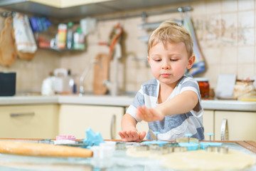 Fototapeta premium Little blond kid plays with molds for making ginger biscuits or cookies, sitting at the kitchen table with raw dough and wheat flour. Rapt face expression. Looking ahead.