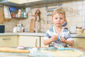 Fototapeta premium Małe blond dziecko bawi się foremkami do robienia ciastek imbirowych lub ciastek, siedząc przy kuchennym stole z surowym ciastem i mąką pszenną. Zachwycony wyraz twarzy. Patrząc w przyszłość.